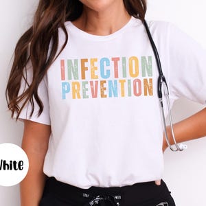 May include: White t-shirt with the words "INFECTION PREVENTION" in colorful, textured letters. A stethoscope hangs around the neck. The shirt is a crew neck style.