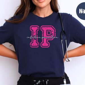 Puede incluir: Camiseta azul marino con las letras "IP" en fuente rosa desgastada, con las palabras "Infection Prevention" en fuente cursiva. Un estetoscopio cuelga alrededor del cuello.