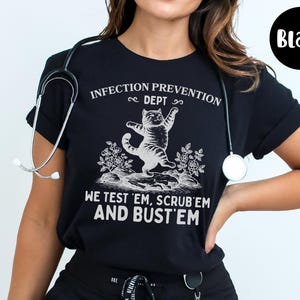 May include: Black t-shirt with a white graphic of a cat and the text "INFECTION PREVENTION DEPT, WE TEST 'EM, SCRUB 'EM AND BUST 'EM". A stethoscope is around the neck.