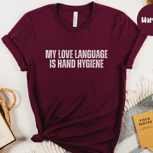 May include: Maroon t-shirt with the text "MY LOVE LANGUAGE IS HAND HYGIENE" in white. The shirt is a solid color and has a crew neck. The sleeves are rolled up. The shirt is made of a soft material.