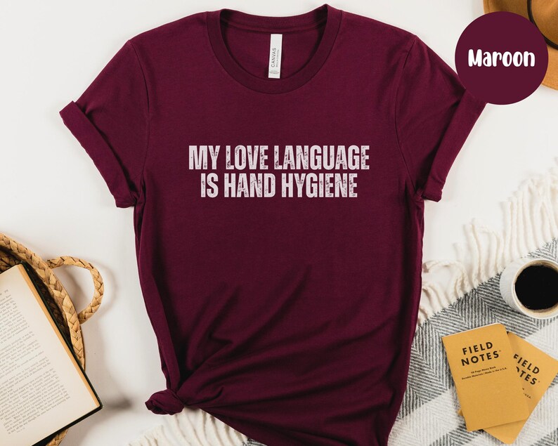 May include: Maroon t-shirt with the text "MY LOVE LANGUAGE IS HAND HYGIENE" in white. The shirt is a solid color and has a crew neck. The sleeves are rolled up. The shirt is made of a soft material.