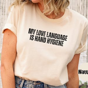 May include: A natural-colored t-shirt with the text "MY LOVE LANGUAGE IS HAND HYGIENE" in black block letters. The shirt has short sleeves and is made of a soft material. The shirt is a casual wear item.