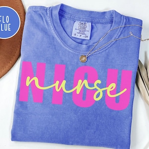 May include: A blue Comfort Colors t-shirt with the words "NICU nurse" in bright pink and yellow script. A gold necklace with a pendant rests on the shirt. The shirt is folded and displayed on a white surface.