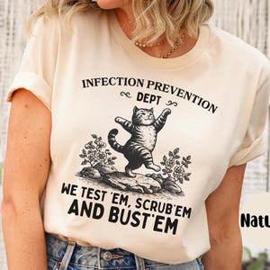 May include: Cream-colored t-shirt with a black and white illustration of a cat standing on its hind legs. The text reads "INFECTION PREVENTION DEPT" and "WE TEST 'EM, SCRUB 'EM AND BUST 'EM". The word "Natural" is also printed on the shirt.