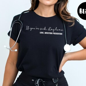 Puede incluir: Camiseta negra con el texto "If you're sick, stay home. LOVE, INFECTION PREVENTION." Un estetoscopio est&aacute; alrededor del cuello. Un c&iacute;rculo negro con la palabra "Black" est&aacute; en la esquina superior derecha.
