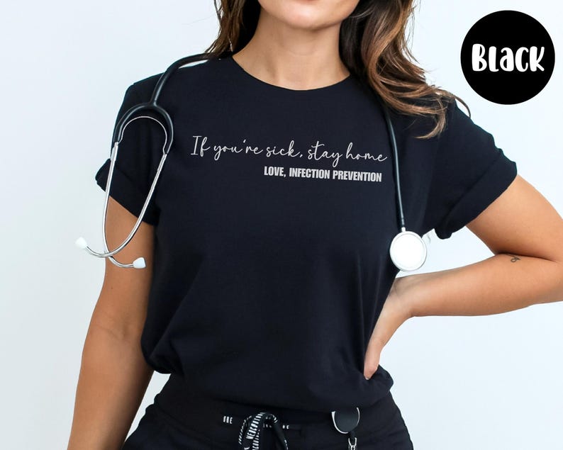 Puede incluir: Camiseta negra con el texto "If you're sick, stay home. LOVE, INFECTION PREVENTION." Un estetoscopio est&aacute; alrededor del cuello. Un c&iacute;rculo negro con la palabra "Black" est&aacute; en la esquina superior derecha.