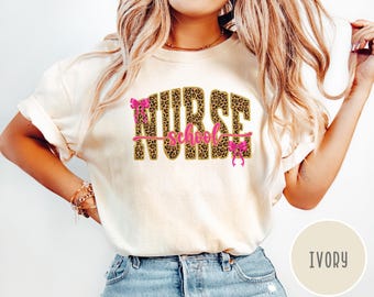 Pode incluir: Uma t-shirt de manga curta na cor marfim com o texto NURSE num padrão de leopardo castanho e preto. A palavra school está escrita em letra cursiva rosa no centro. Dois pequenos laços cor-de-rosa estão presos às letras N e E.