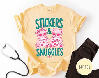 Puede incluir: Una camiseta de color amarillo pálido con un gráfico de dos ositos de peluche rosas con gafas de sol en forma de corazón. Los osos sostienen hojas de pegatinas. El texto dice STICKERS & SNUGGLES en turquesa. Un estetoscopio es visible.
