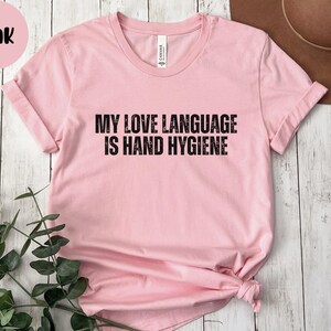 May include: A light pink t-shirt with the text "MY LOVE LANGUAGE IS HAND HYGIENE" in black block letters. The shirt has short sleeves and a crew neck. A pink circle with the word "Pink" is in the upper left corner.
