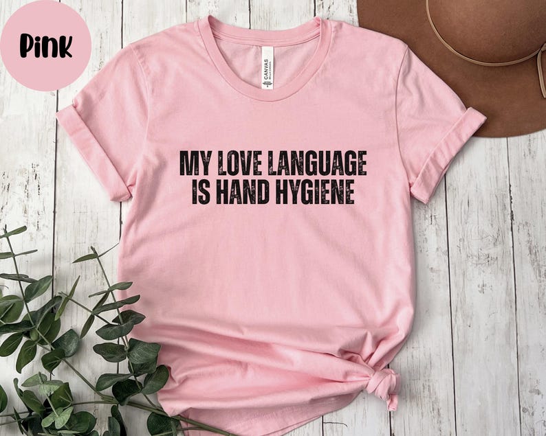 May include: A light pink t-shirt with the text "MY LOVE LANGUAGE IS HAND HYGIENE" in black block letters. The shirt has short sleeves and a crew neck. A pink circle with the word "Pink" is in the upper left corner.