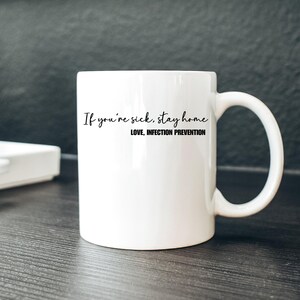 Puede incluir: Taza de cer&aacute;mica blanca con asa curva. La taza presenta el texto "If you're sick, stay home" en estilo manuscrito, con "LOVE, INFECTION PREVENTION" debajo. La taza est&aacute; sobre una superficie oscura.