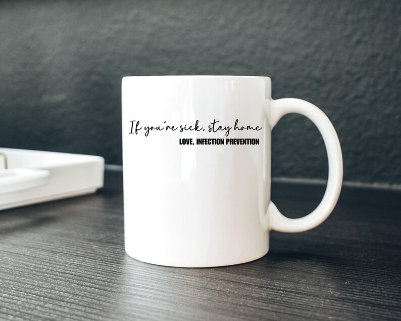 Puede incluir: Taza de cer&aacute;mica blanca con asa curva. La taza presenta el texto "If you're sick, stay home" en estilo manuscrito, con "LOVE, INFECTION PREVENTION" debajo. La taza est&aacute; sobre una superficie oscura.