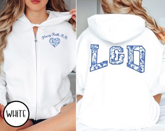 May include: White zip-up hoodie with blue floral design. Features "Mary Ruth R.N." and a heart on the front. The back displays "LGD" in a blue floral pattern. A casual, comfortable garment.