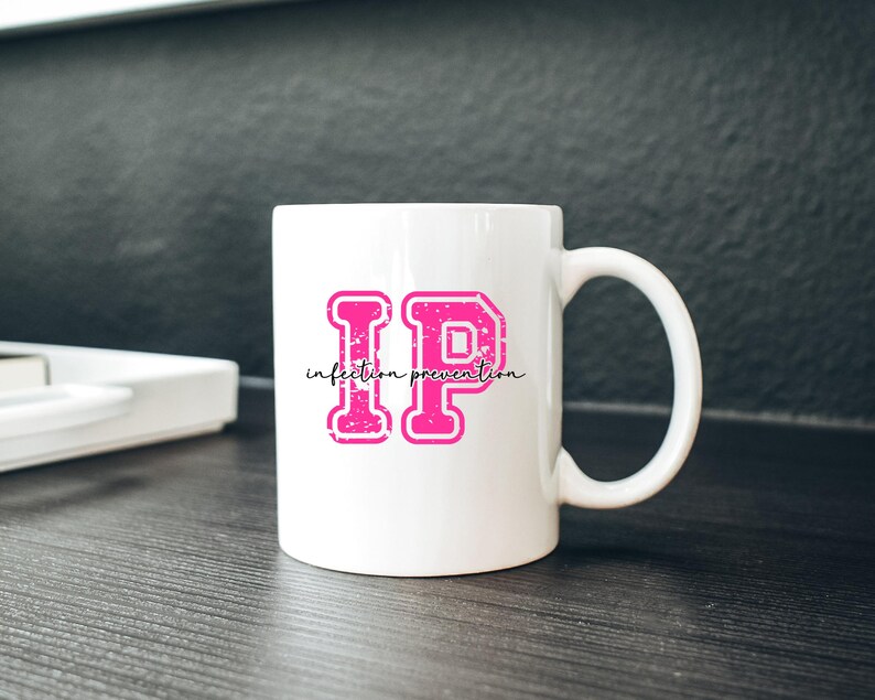 Puede incluir: Taza de cer&aacute;mica blanca con un gran dise&ntilde;o "IP" rosa y las palabras "infection prevention" en una fuente cursiva. Las letras tienen un aspecto texturizado y desgastado. La taza tiene un asa curva y est&aacute; sobre una superficie oscura.
