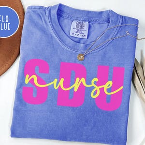 May include: A blue t-shirt with the text "SDU nurse" in pink and yellow. The shirt is folded and has a Comfort Colors tag. A gold necklace with a pendant is draped across the shirt.