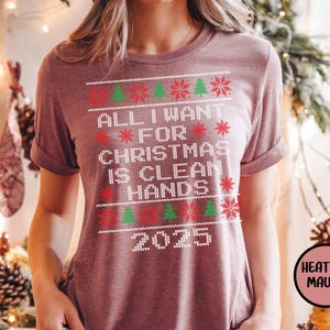 May include: Heather mauve t-shirt with the text "ALL I WANT FOR CHRISTMAS IS CLEAN HANDS 2025" in white. The design includes red snowflakes and green Christmas trees. The shirt has short sleeves.