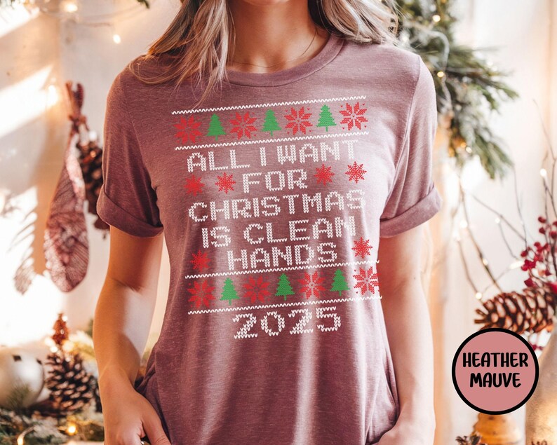 May include: Heather mauve t-shirt with the text "ALL I WANT FOR CHRISTMAS IS CLEAN HANDS 2025" in white. The design includes red snowflakes and green Christmas trees. The shirt has short sleeves.