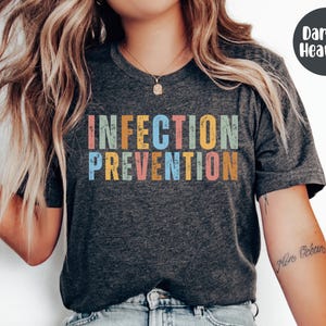 May include: Dark gray heather t-shirt with the words "INFECTION PREVENTION" in colorful, distressed lettering. The text is stacked in two lines, with each word in a different color. A dark gray circle with the words "Dark G. Heather" is in the upper right corner.