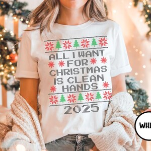 May include: White t-shirt with a festive design. The shirt features the text "ALL I WANT FOR CHRISTMAS IS CLEAN HANDS 2025" in a pixelated, sweater-like font, with red snowflakes and green Christmas trees. The word "WHITE" is in a black circle.