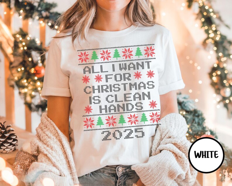May include: White t-shirt with a festive design. The shirt features the text "ALL I WANT FOR CHRISTMAS IS CLEAN HANDS 2025" in a pixelated, sweater-like font, with red snowflakes and green Christmas trees. The word "WHITE" is in a black circle.