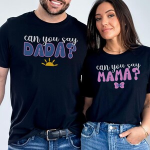 May include: Two black t-shirts with a gender reveal announcement. The shirt on the left says "Can you say DADA?" with a yellow sun graphic. The shirt on the right says "Can you say MAMA?" with a pink bow graphic.