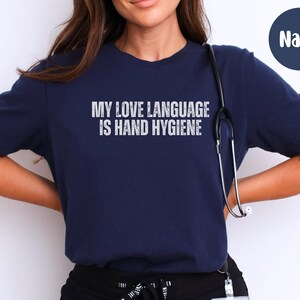 May include: Navy blue t-shirt with the text "MY LOVE LANGUAGE IS HAND HYGIENE" in white, distressed lettering. A stethoscope hangs around the neck. The shirt is a crew neck style.