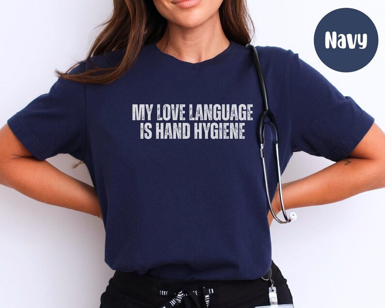 May include: Navy blue t-shirt with the text "MY LOVE LANGUAGE IS HAND HYGIENE" in white, distressed lettering. A stethoscope hangs around the neck. The shirt is a crew neck style.