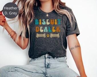 May include: A heather grey t-shirt with the text "BISCUIT DEALER" in colourful retro font. The text is surrounded by two bone-shaped dog biscuits and three paw prints.