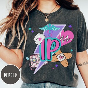 May include: Dark gray t-shirt featuring a colorful graphic design. The design includes a nurse's hat, stethoscope, heart, bandages, and the letters "IP" in a stylized font. The shirt has a vintage, washed look.