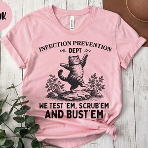 May include: Pink t-shirt with the text "INFECTION PREVENTION DEPT" and "WE TEST 'EM, SCRUB 'EM AND BUST 'EM". The shirt features a graphic of a cat standing on a rock. The shirt is made of a soft material.