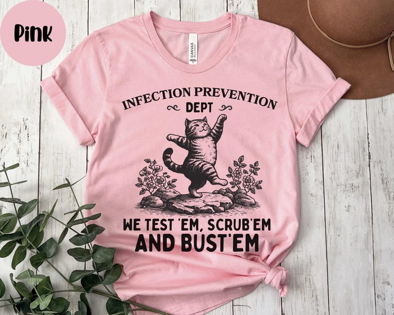 May include: Pink t-shirt with the text "INFECTION PREVENTION DEPT" and "WE TEST 'EM, SCRUB 'EM AND BUST 'EM". The shirt features a graphic of a cat standing on a rock. The shirt is made of a soft material.