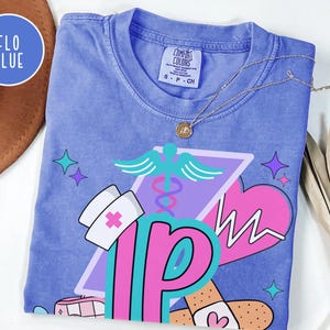 May include: A blue Comfort Colors t-shirt with a graphic design featuring a nurse's hat, a caduceus, a heart with a heartbeat line, and the letters "NP" in pink and teal. The shirt has a crew neck and short sleeves. The text "FLO BLUE" is in a circle.