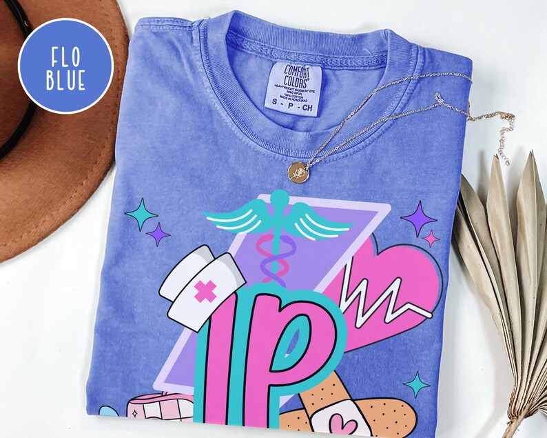 May include: A blue Comfort Colors t-shirt with a graphic design featuring a nurse's hat, a caduceus, a heart with a heartbeat line, and the letters "NP" in pink and teal. The shirt has a crew neck and short sleeves. The text "FLO BLUE" is in a circle.