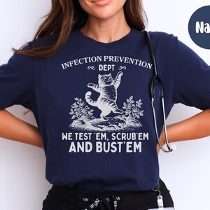 May include: Navy blue t-shirt with white text and a graphic of a cat. The text reads "INFECTION PREVENTION DEPT" above the cat, and "WE TEST 'EM, SCRUB 'EM AND BUST 'EM" below. A stethoscope hangs on the shirt.