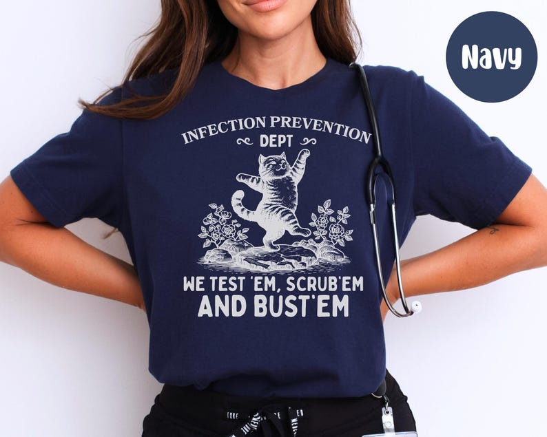 May include: Navy blue t-shirt with white text and a graphic of a cat. The text reads "INFECTION PREVENTION DEPT" above the cat, and "WE TEST 'EM, SCRUB 'EM AND BUST 'EM" below. A stethoscope hangs on the shirt.