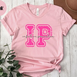 Puede incluir: Camiseta rosa con las letras "IP" en una fuente desgastada de estilo universitario, con las palabras "infection prevention" en cursiva debajo. Un c&iacute;rculo rosa con la palabra "Pink" est&aacute; en la esquina superior izquierda.