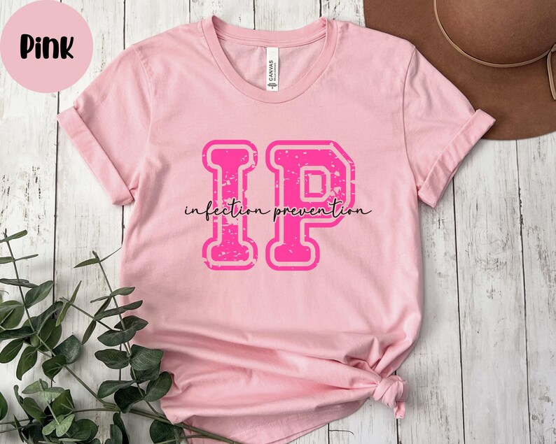 Puede incluir: Camiseta rosa con las letras "IP" en una fuente desgastada de estilo universitario, con las palabras "infection prevention" en cursiva debajo. Un c&iacute;rculo rosa con la palabra "Pink" est&aacute; en la esquina superior izquierda.