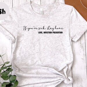 Puede incluir: Camiseta gris claro con el texto "If you're sick, stay home. LOVE, INFECTION PREVENTION" en escritura negra. La camiseta est&aacute; anudada en la parte inferior. La palabra "Ash" est&aacute; en un c&iacute;rculo en la esquina superior izquierda.