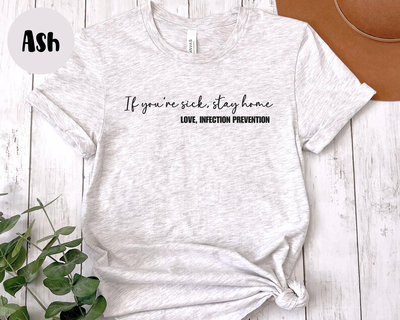Puede incluir: Camiseta gris claro con el texto "If you're sick, stay home. LOVE, INFECTION PREVENTION" en escritura negra. La camiseta est&aacute; anudada en la parte inferior. La palabra "Ash" est&aacute; en un c&iacute;rculo en la esquina superior izquierda.