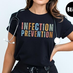 May include: Black t-shirt with the words "INFECTION PREVENTION" in colorful, distressed lettering. A stethoscope is draped around the neck. A black circle with the word "Black" is in the upper right corner.