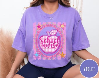 Pode incluir: Uma t-shirt violeta de manga curta com um gráfico central de uma maçã aos quadrados cor-de-rosa e brancos com o texto "School nurse" em letra cursiva. O design é emoldurado por um círculo recortado, riscas cor-de-rosa e quatro estrelas coloridas.