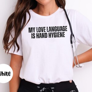 May include: White t-shirt with the text "MY LOVE LANGUAGE IS HAND HYGIENE" in black. A stethoscope hangs around the neck. The shirt is a crew neck style.
