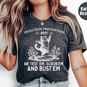 May include: Dark heather gray t-shirt with the text "INFECTION PREVENTION DEPT" and "WE TEST 'EM, SCRUB 'EM AND BUST 'EM". The shirt features a white graphic of a cat standing on its hind legs. The shirt is made of a soft material.