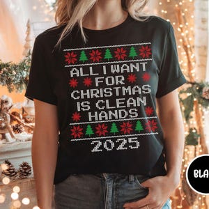May include: Black t-shirt with a festive Christmas design. The shirt features the text "ALL I WANT FOR CHRISTMAS IS CLEAN HANDS 2025" in white, red, and green. The design includes Christmas trees and snowflakes.