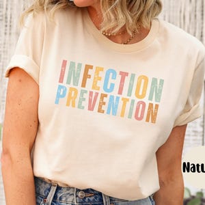 May include: A cream-colored t-shirt with the words "INFECTION PREVENTION" printed in colorful, distressed lettering. The shirt has short sleeves and a crew neck. The word "Natural" is printed on a white circle.