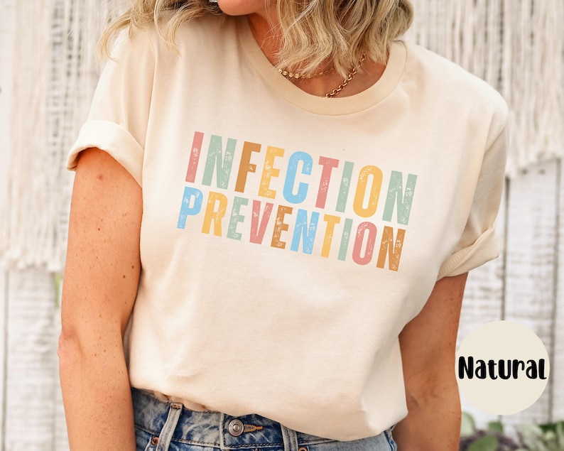 May include: A cream-colored t-shirt with the words "INFECTION PREVENTION" printed in colorful, distressed lettering. The shirt has short sleeves and a crew neck. The word "Natural" is printed on a white circle.
