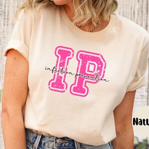 Puede incluir: Una camiseta color crema con las letras "IP" en rosa intenso y en negrita, con un estilo universitario. Las palabras "infection prevention" est&aacute;n escritas en cursiva debajo. La camiseta est&aacute; etiquetada como "Natural".