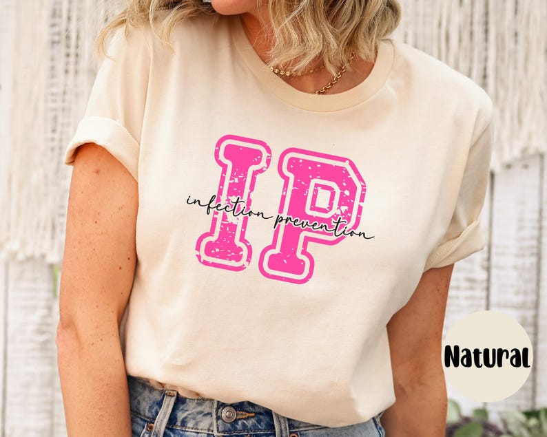 Puede incluir: Una camiseta color crema con las letras "IP" en rosa intenso y en negrita, con un estilo universitario. Las palabras "infection prevention" est&aacute;n escritas en cursiva debajo. La camiseta est&aacute; etiquetada como "Natural".