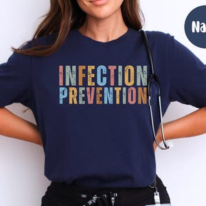 May include: Navy blue t-shirt with the words "INFECTION PREVENTION" in colorful, vintage-style lettering. A stethoscope hangs around the neck. The shirt is a crew neck and has short sleeves.