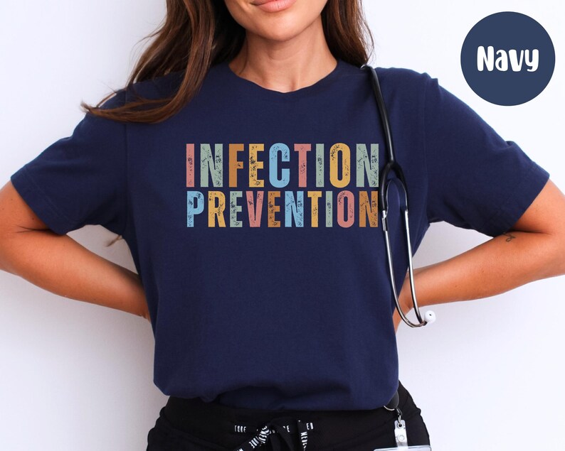 May include: Navy blue t-shirt with the words "INFECTION PREVENTION" in colorful, vintage-style lettering. A stethoscope hangs around the neck. The shirt is a crew neck and has short sleeves.